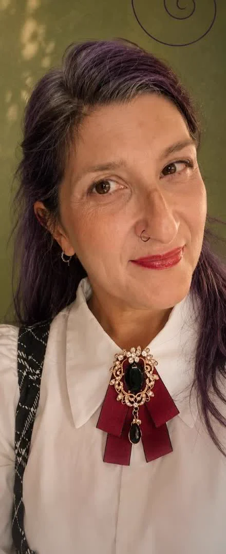 Mariangela, capelli viola e camicia bianca con una spilla, sorride davanti a uno sfondo verde oliva